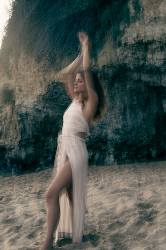 Natalie Montoya moving freely in a white dress in front of a rock face on the beach